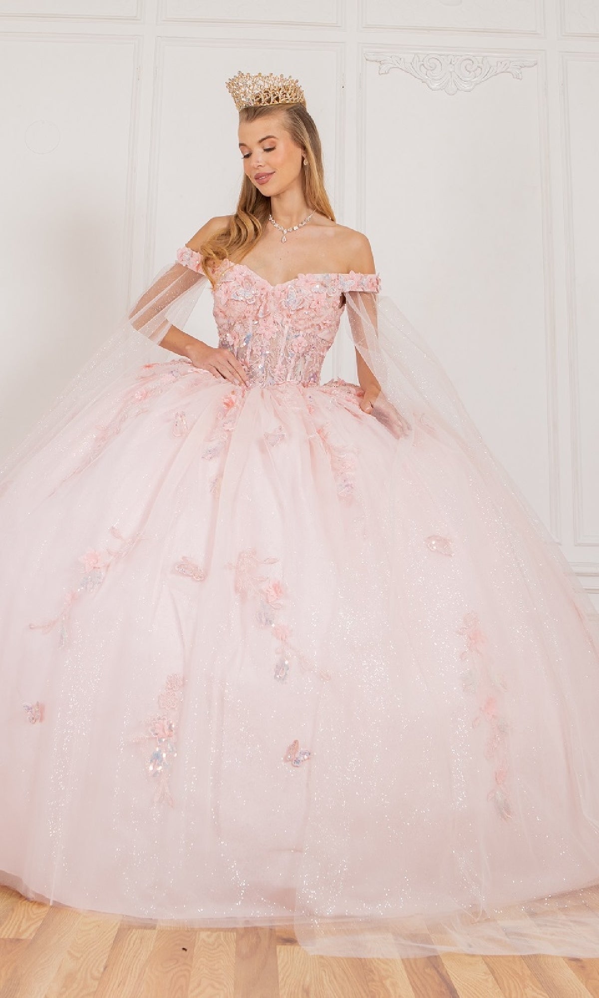 A woman stands indoors on a wooden floor wearing the Cinderella Couture 8075J Quinceanera Dress with sparkly, off-the-shoulder design and floral 3D appliques, accessorized with a jeweled crown and necklace.